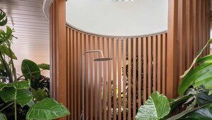 Un jardín interior moderno con una pared de madera curva, una fuente de agua metálica y abundante vegetación verde.