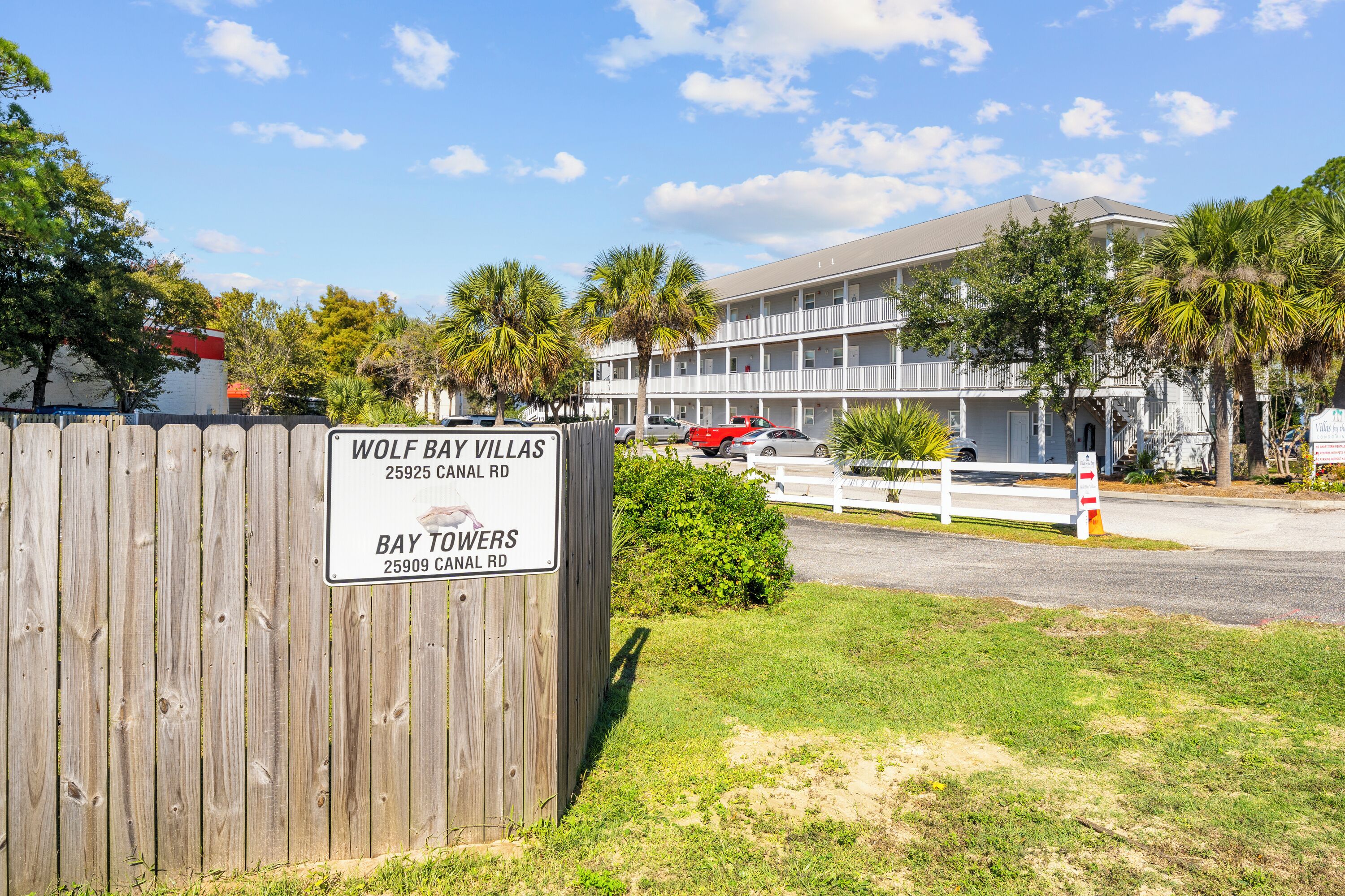 Een houten hek met borden voor Wolf Bay Villas en Bay Towers, een meer verdiepingen tellend gebouw met balkons, en een heldere lucht.