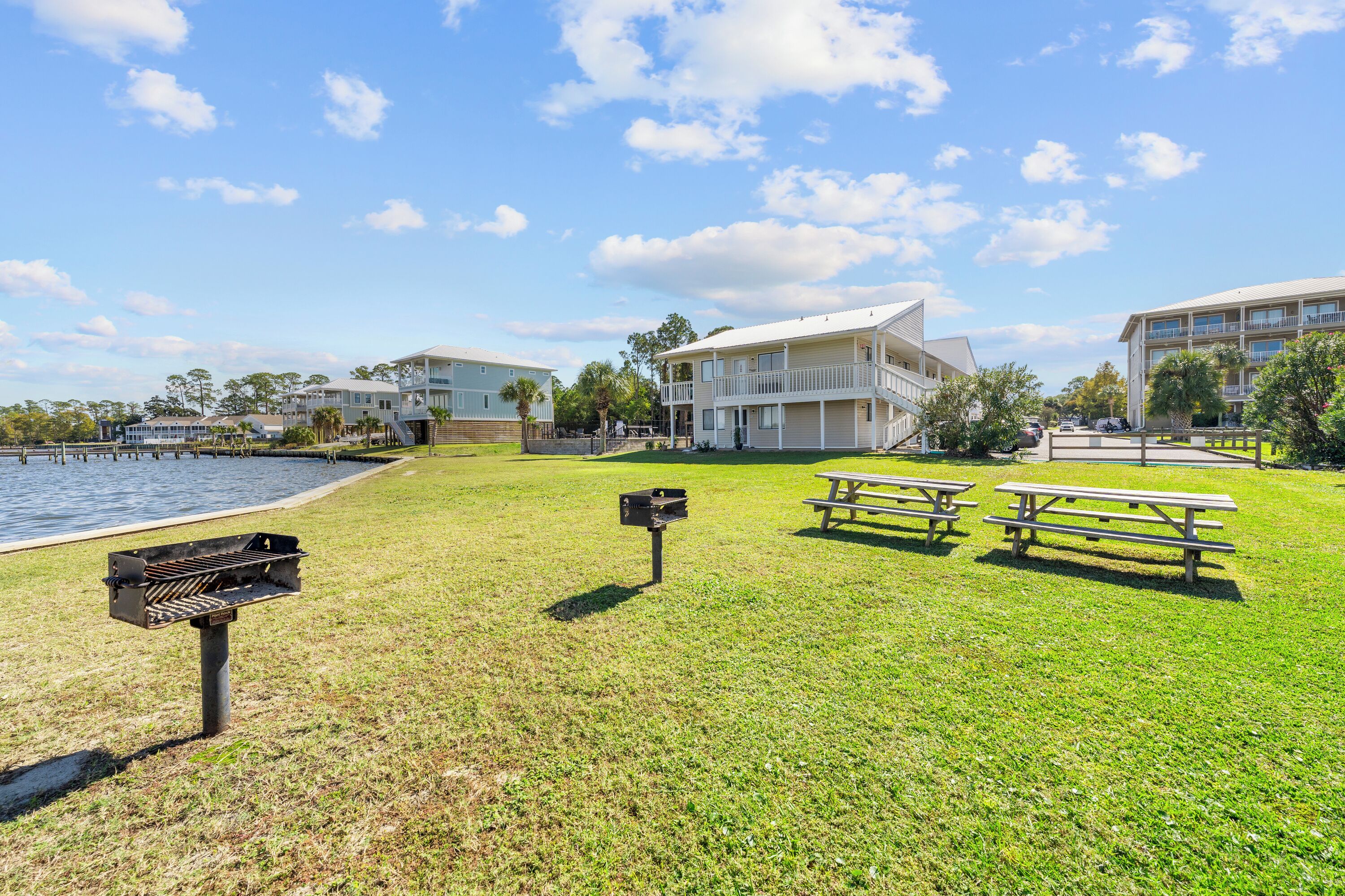 A grassy area with picnic tables and a barbecue grill, overlooking a body of water and buildings in the background.
