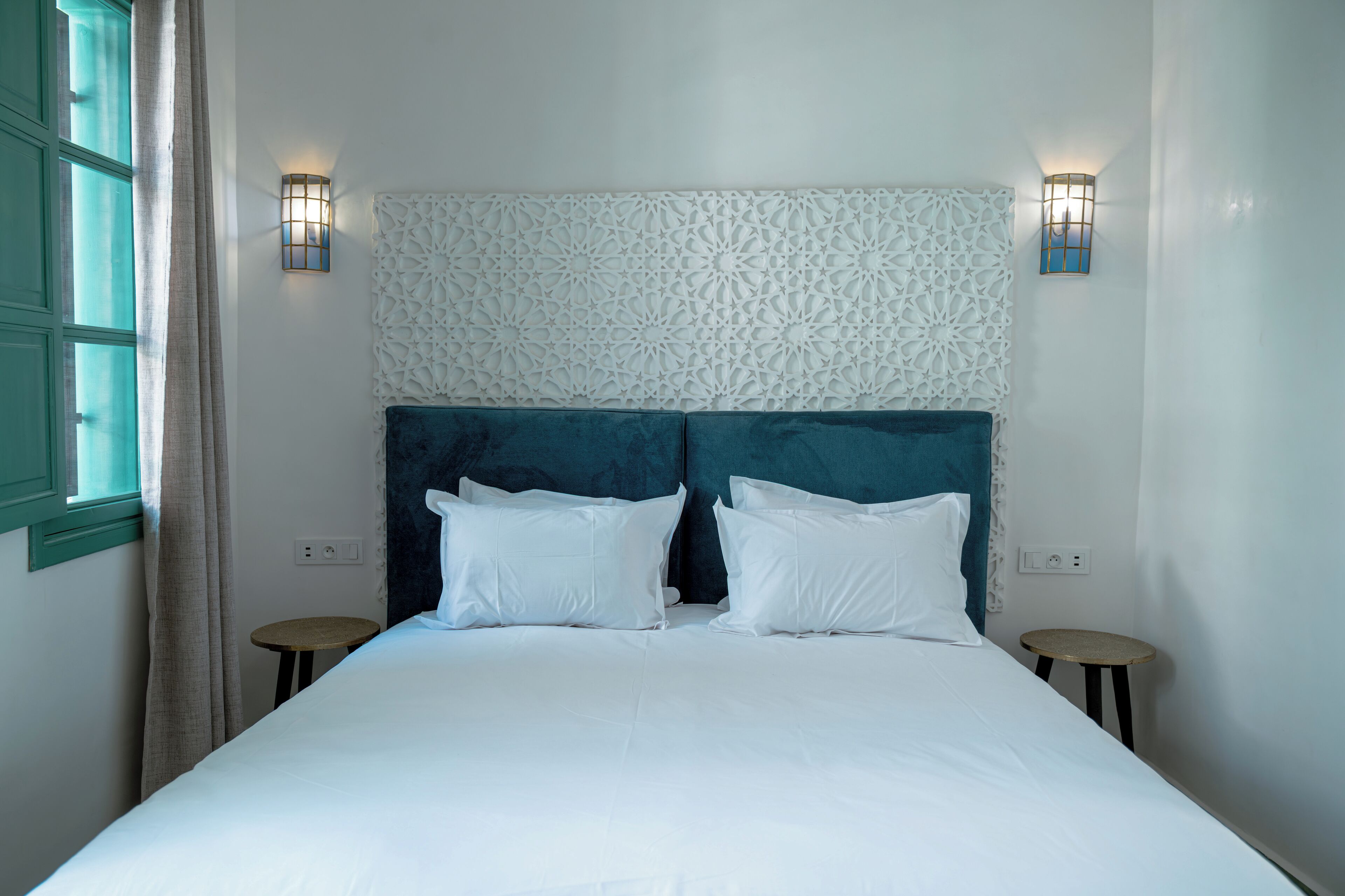Una cama bien hecha con sábanas blancas y un cabecero verde agua, dos apliques de pared y una mesita redonda al lado de la cama.