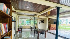 A spacious indoor area with a wooden ceiling, a dining table with chairs, and a bookshelf filled with books. - Daosri The Inn (Barangabari)