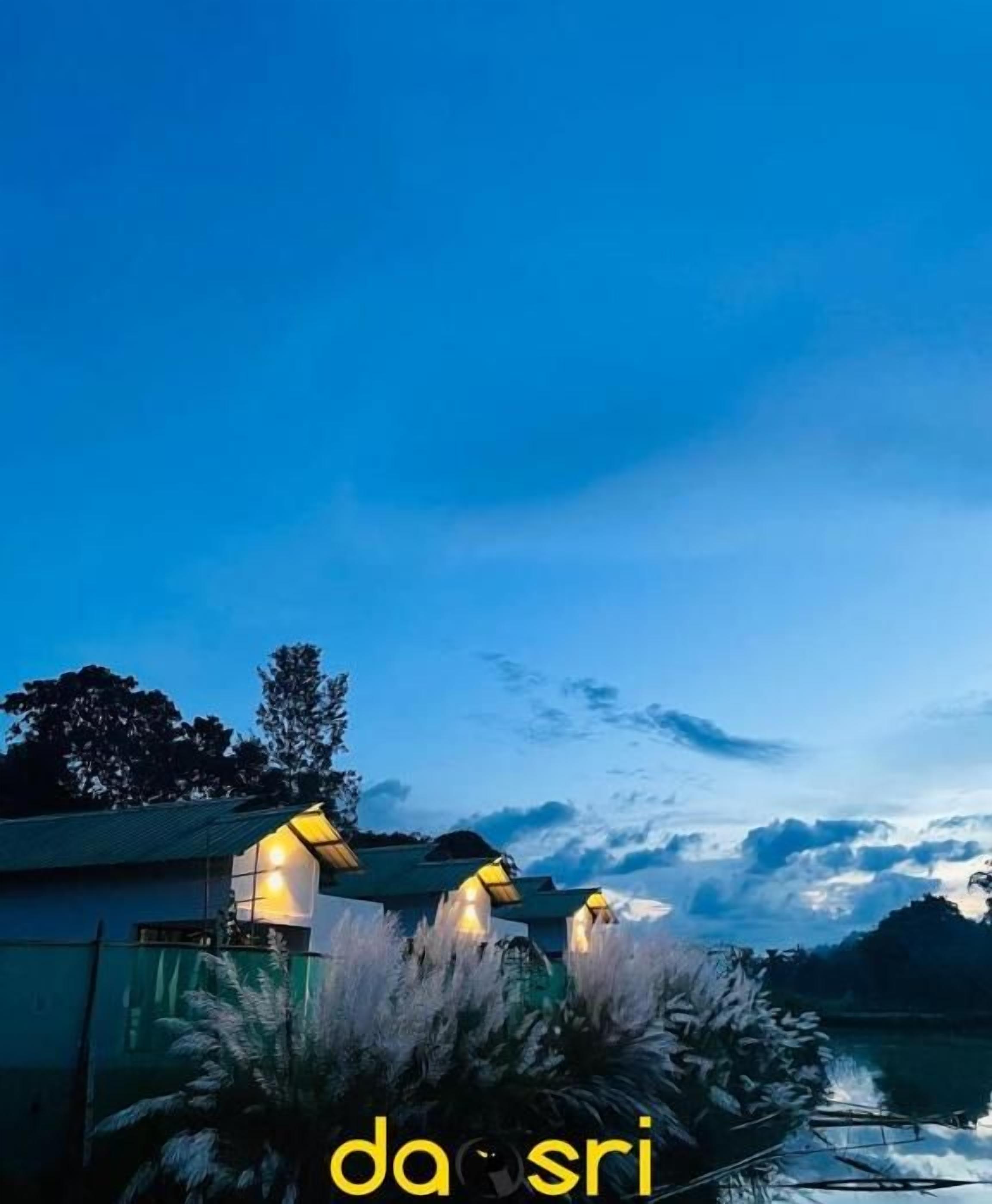 A serene lakeside view at dusk with two illuminated structures, possibly a gazebo or pavilion, surrounded by lush vegetation.