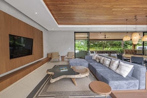 Una sala de estar moderna con un televisor de pantalla plana grande, un sofá gris con cojines, una mesa de centro original y una zona de comedor con asientos exteriores.