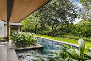 A modern outdoor pool area with a wooden pergola, surrounded by lush greenery and a well-maintained lawn.