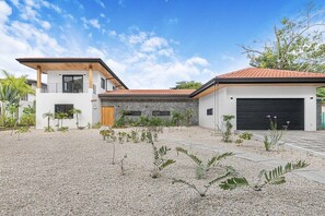 A modern two-story house with a gravel driveway, a garage, and a small garden with various plants. - Casa Campana (Santa Cruz)