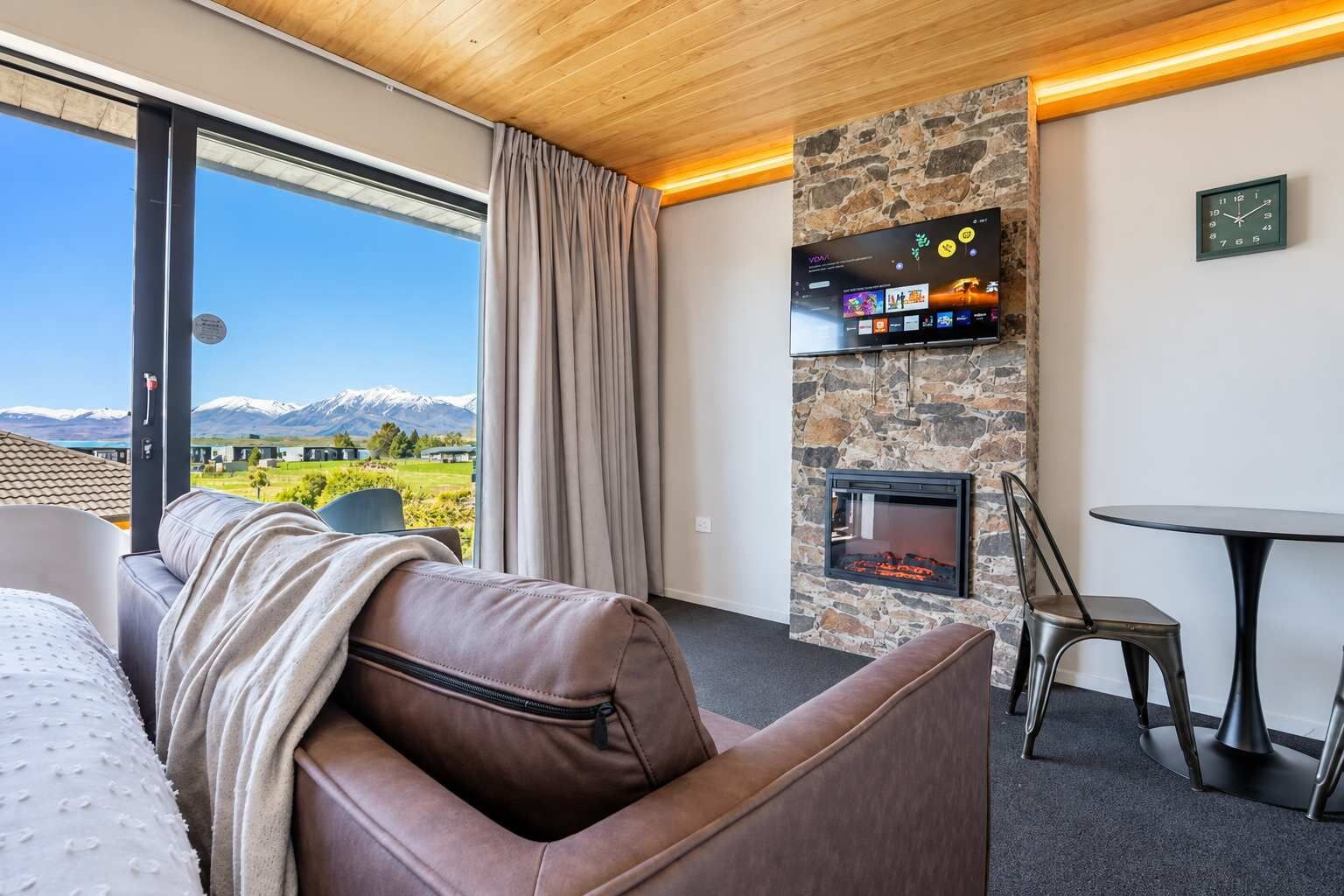 Habitación de hotel con sofá de cuero, chimenea de piedra, televisión de pantalla plana y vistas a un paisaje montañoso.
