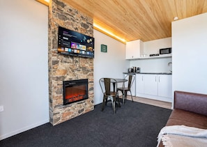 A modern living area with a stone fireplace, a flat-screen TV mounted on the wall, a dining table with chairs, and a kitchenette in the background. - Blue Sky | Lake Views + Private Deck + Stargazing (Lake Tekapo)