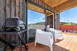 Une terrasse extérieure moderne, dotée d’un barbecue, de deux chaises longues blanches et d’une porte vitrée donnant sur un espace abrité.