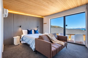 A modern bedroom with a large bed, a brown leather sofa, and a view of the mountains through a large window. - Blue Sky | Lake Views + Private Deck + Stargazing (Lake Tekapo)