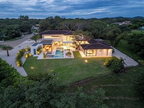 Ein modernes, zweistöckiges Haus mit Swimmingpool, umgeben von üppiger Vegetation und Hügeln im Hintergrund.
