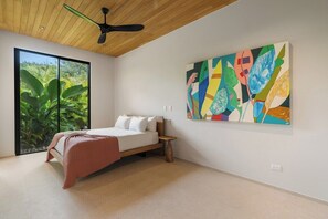 A bedroom with a bed, a ceiling fan, a large window with a view of greenery, and a colorful abstract painting on the wall. - Lingalonga (Guanacaste)