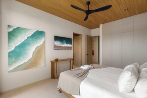 A bedroom with a bed, a ceiling fan, a painting of a wave, and a wooden console table. - Lingalonga (Guanacaste)