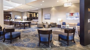 Un vestíbulo de hotel espacioso con un techo alto, una zona de estar central con sofás y sillones, una mesa de comedor y una escalera que conduce a otro nivel.