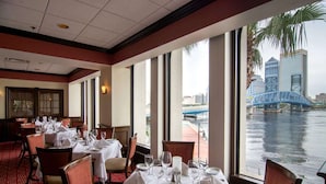 Un comedor con mesas dispuestas para el servicio, amplias ventanas con vistas a un puente y al agua, y un suelo cubierto de alfombra roja.