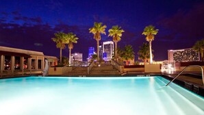 Une piscine illuminée la nuit, avec la silhouette de la ville en arrière-plan, des palmiers et un escalier menant à un niveau supérieur.