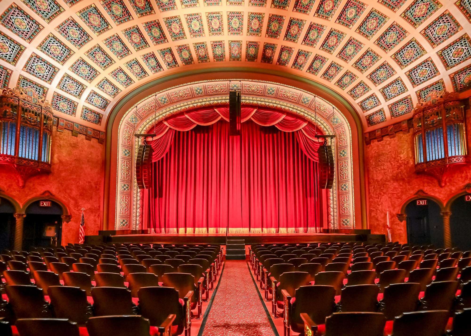 An auditorium with rows of seats, a red curtain backdrop, and a patterned ceiling.