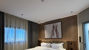 Une chambre d’hôtel moderne avec un grand lit, une tête de lit en bois, un tableau au-dessus du lit et une vue sur la ville par la fenêtre.