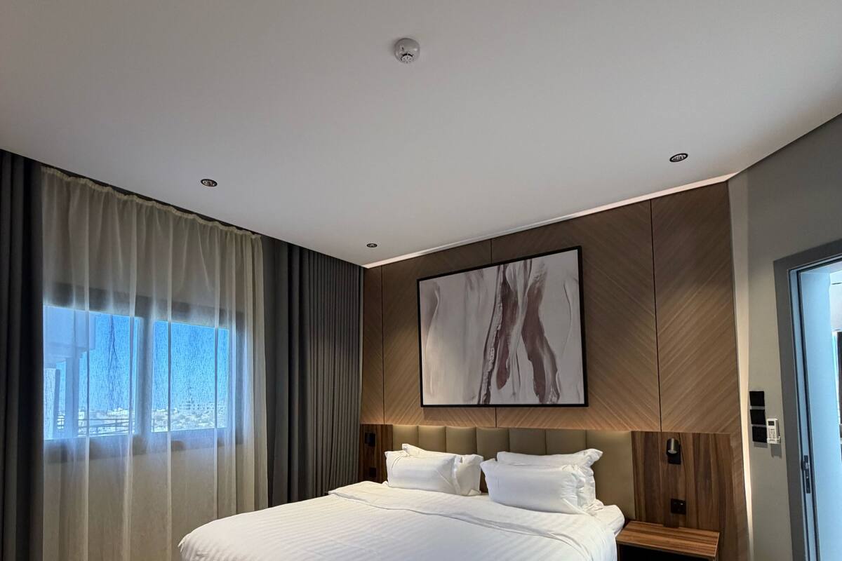 A modern hotel room with a large bed, wooden headboard, a painting above the bed, and a view of the city through the window.