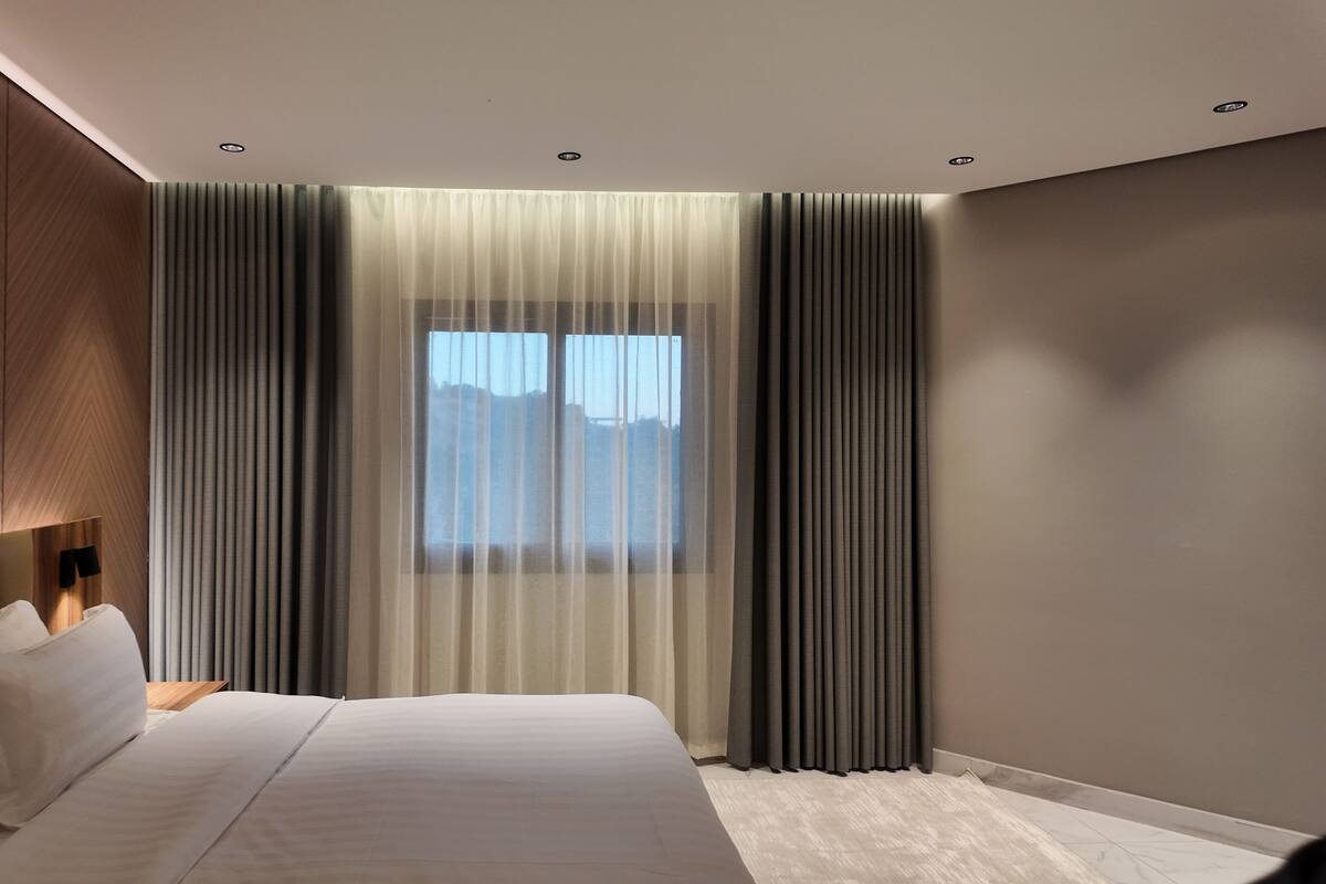 A modern hotel room with a large bed, a headboard, a bedside table, and a window with sheer curtains.