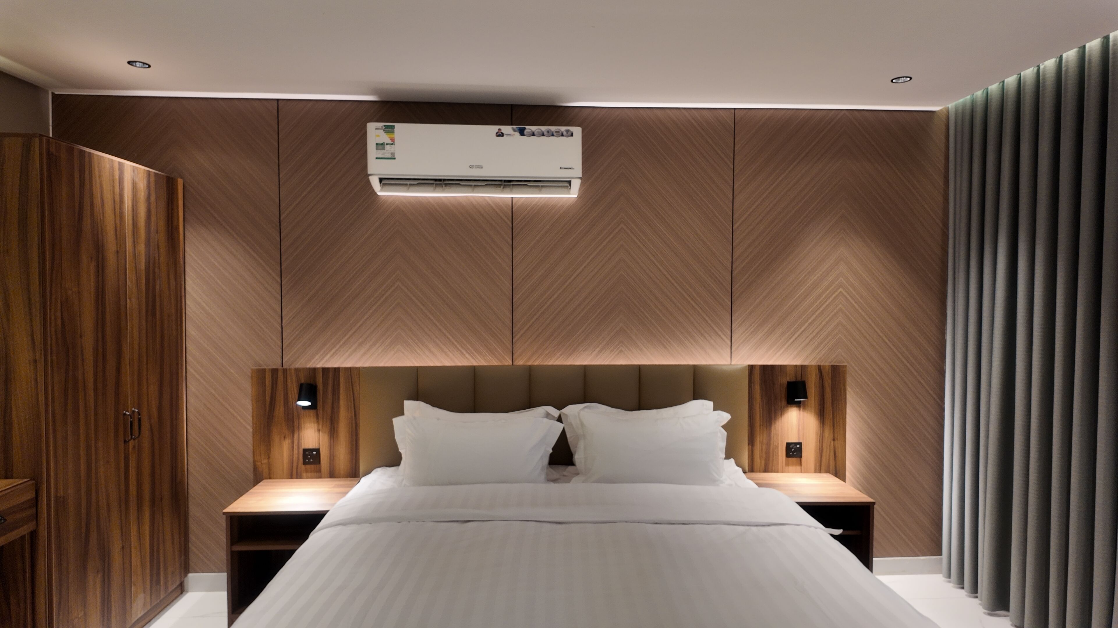 Ein modernes Schlafzimmer mit einem großen Bett, Holzpaneelen, einer Klimaanlage, Nachttischlampen und einem Kleiderschrank.