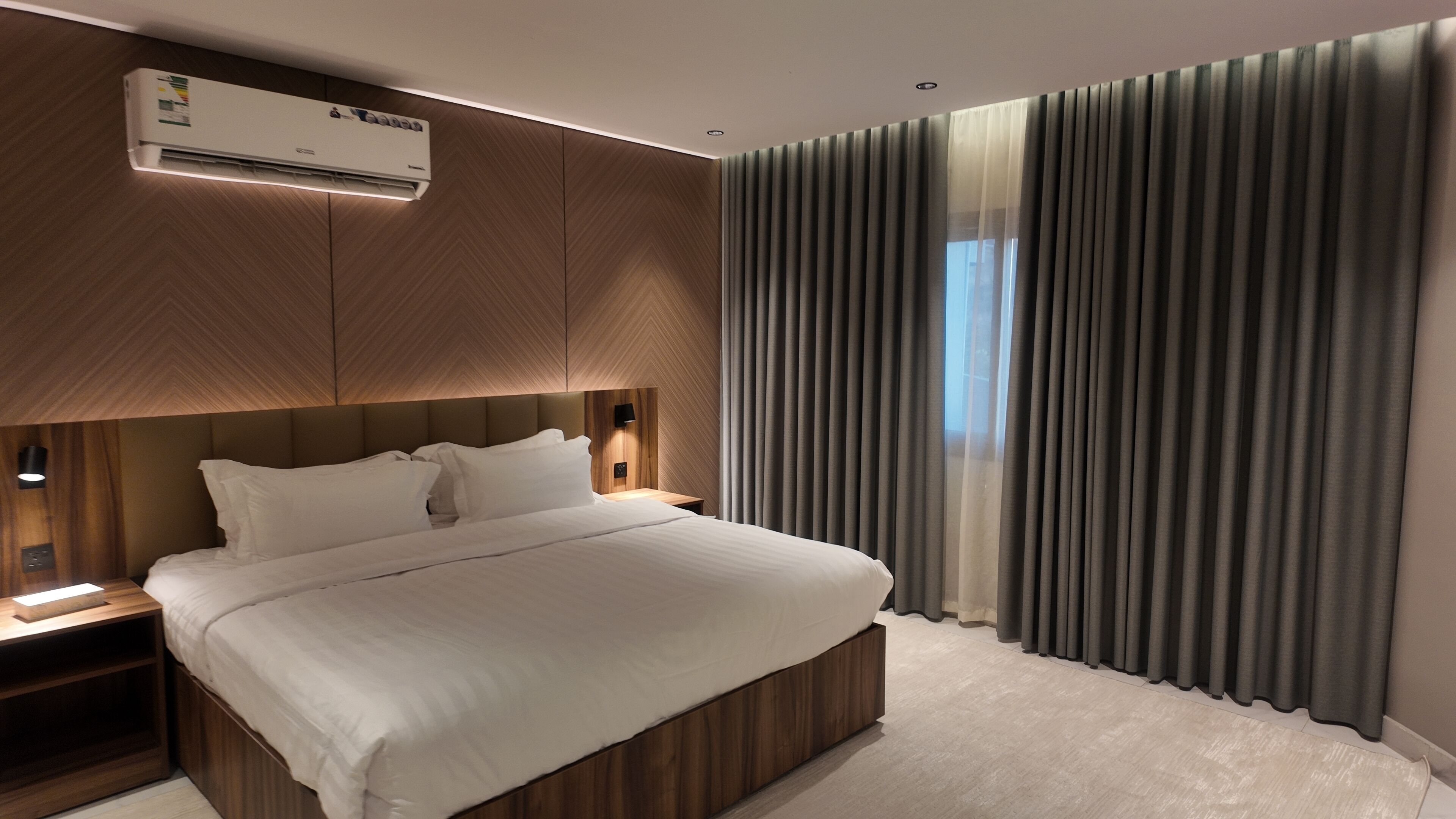 Una habitación de hotel moderna con una cama grande, paneles de madera, aire acondicionado integrado y vistas al exterior a través de cortinas translúcidas.