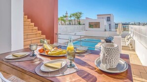 A dining table with plates, cutlery, and a bottle, overlooking a swimming pool and a building with a red wall and stairs. - Casa Bayview Bliss - Sea Views, Pool & BBQ, Stroll to Beach and Ferragudo Village (Ferragudo)