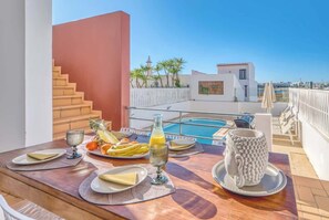 A dining table with plates, cutlery, and a bottle, overlooking a swimming pool and a building with a red wall and stairs.