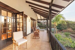 Ein überdachter Balkon mit Blick auf einen Garten, ein Holzschaukelstuhl und ein gepolsterter Stuhl.
