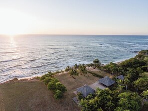 Luftaufnahme eines Küsteneigentums mit Strand, Palmen und strohgedeckten Gebäuden.