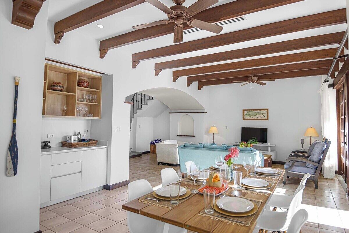 Un espacio habitable amplio con una mesa de comedor preparada para cuatro personas, una cocina con armarios blancos y una escalera que conduce a un nivel superior.
