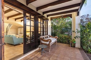 Ein überdachter Patio-Bereich mit einer Bank und einem kleinen Tisch, eine Treppe, die zu einem Innenraum führt, und ein Blick auf einen Garten mit verschiedenen Pflanzen.