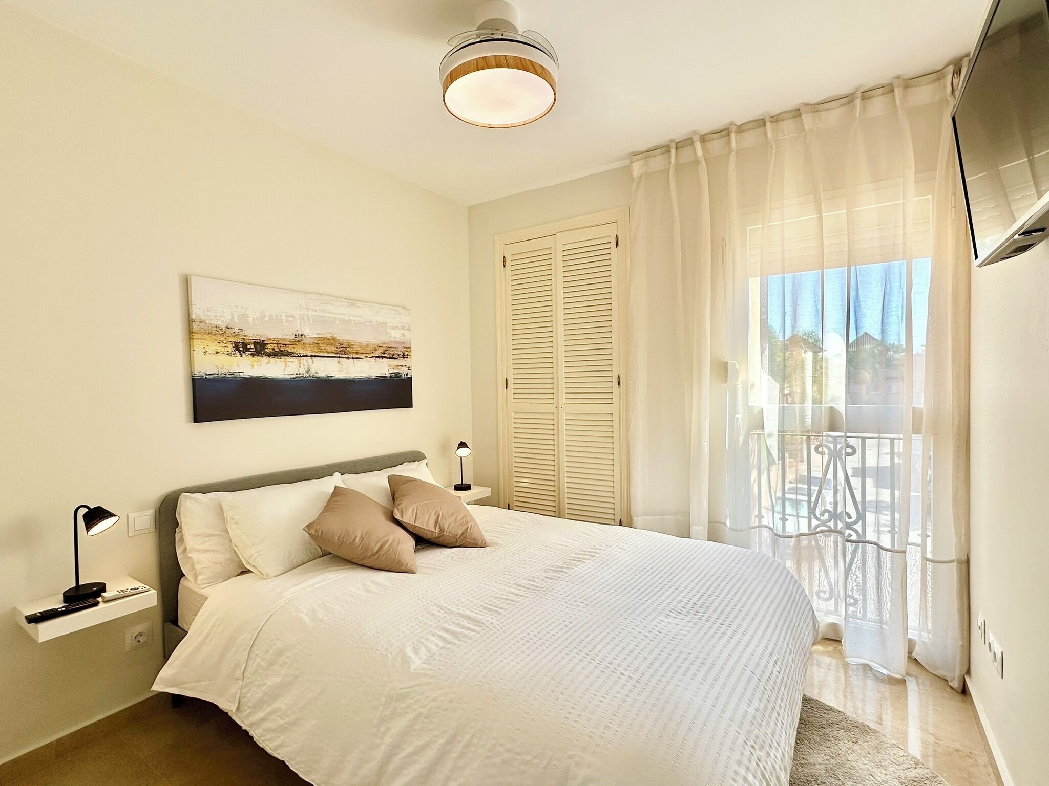 A bedroom with a bed, a nightstand, a lamp, a painting, and a balcony with a view.