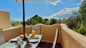 Une table à manger avec des aliments pour le petit-déjeuner, une boisson, et une vue sur un paysage tropical avec des palmiers et des bâtiments.