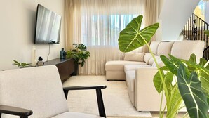 Un salon moderne avec un téléviseur à écran plat, des canapés de couleur beige, une table basse et une grande plante.