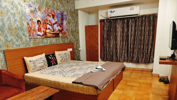 Quarto com cama de madeira, uma pequena mesa de madeira, poltrona vermelha, um quadro na parede e uma janela com cortinas.