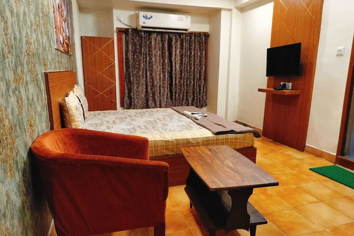 A room with a bed, a red chair, a wooden table, a TV on a wooden shelf, and an air conditioning unit.