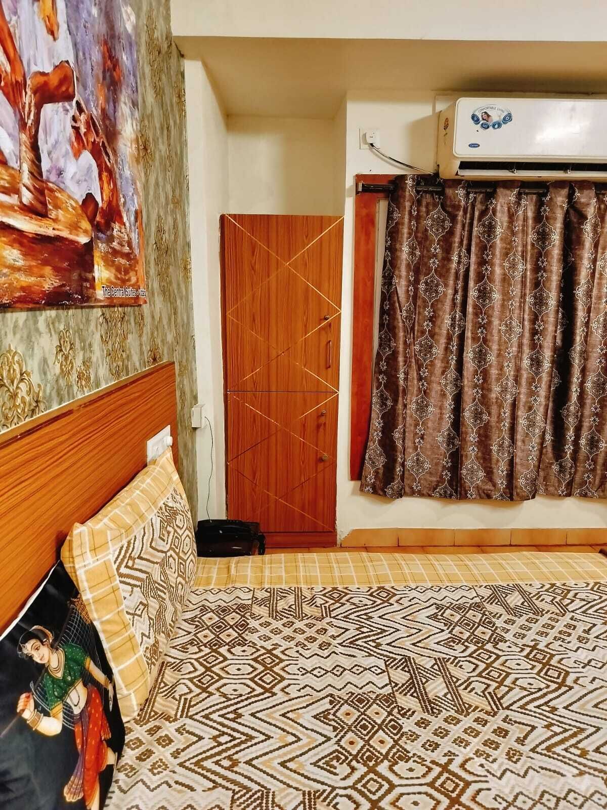 A bedroom with a patterned bedspread, a wooden wardrobe, a painting on the wall, and an air conditioning unit.