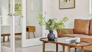 A modern living room with a brown leather sofa, a wooden coffee table with books and a candle, a potted plant, and a framed botanical illustration on the wall. - Sleek Kitchenette & Queen Bed (Fort Lauderdale)