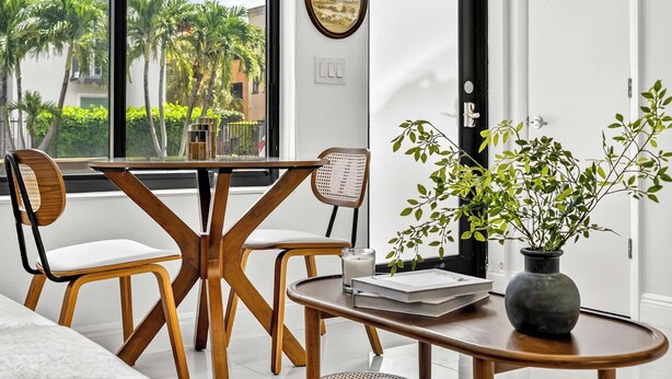 A modern dining area with a wooden table and chairs, a vase with green foliage, and a view of palm trees through a large window. - Sleek Kitchenette & Queen Bed (Fort Lauderdale)