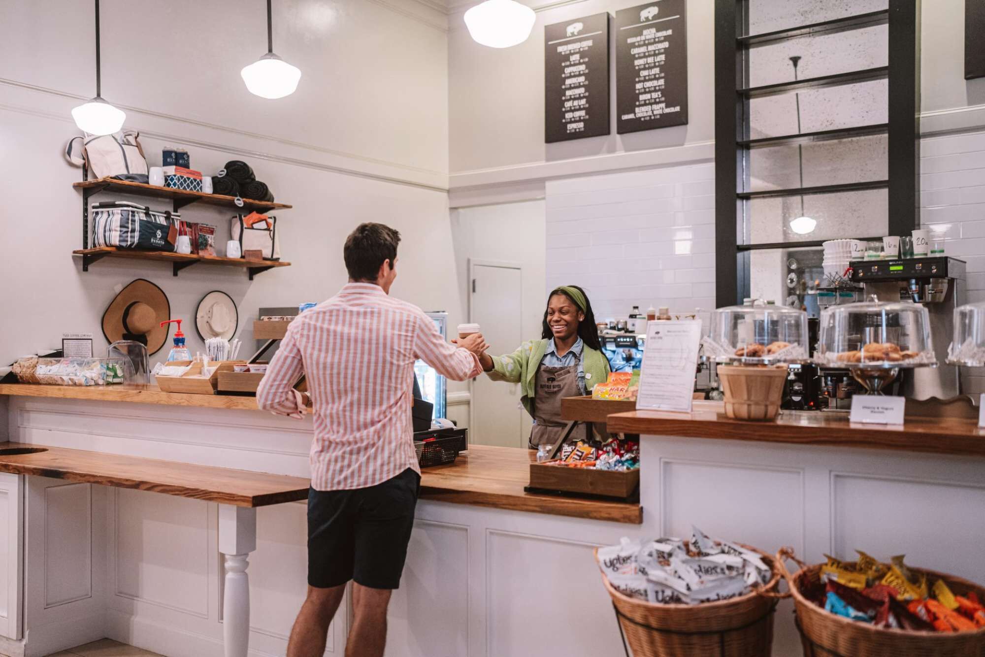 A coffee shop counter with a customer and a barista, a menu board, and baskets of snacks.