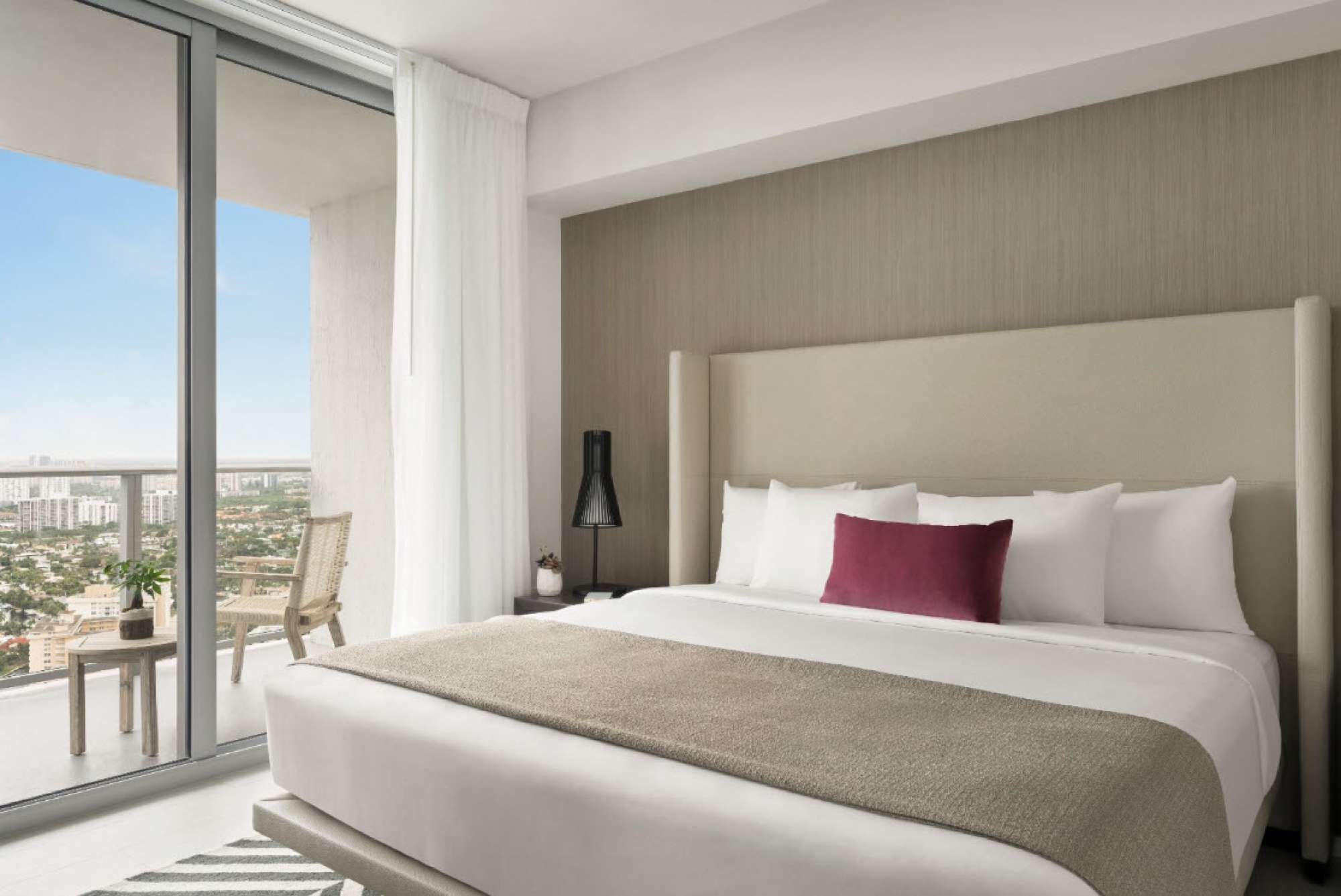 Une chambre d’hôtel moderne, dotée d’un grand lit, d’une table de chevet, d’une lampe et offrant une vue sur la ville grâce à de grandes fenêtres du sol au plafond.