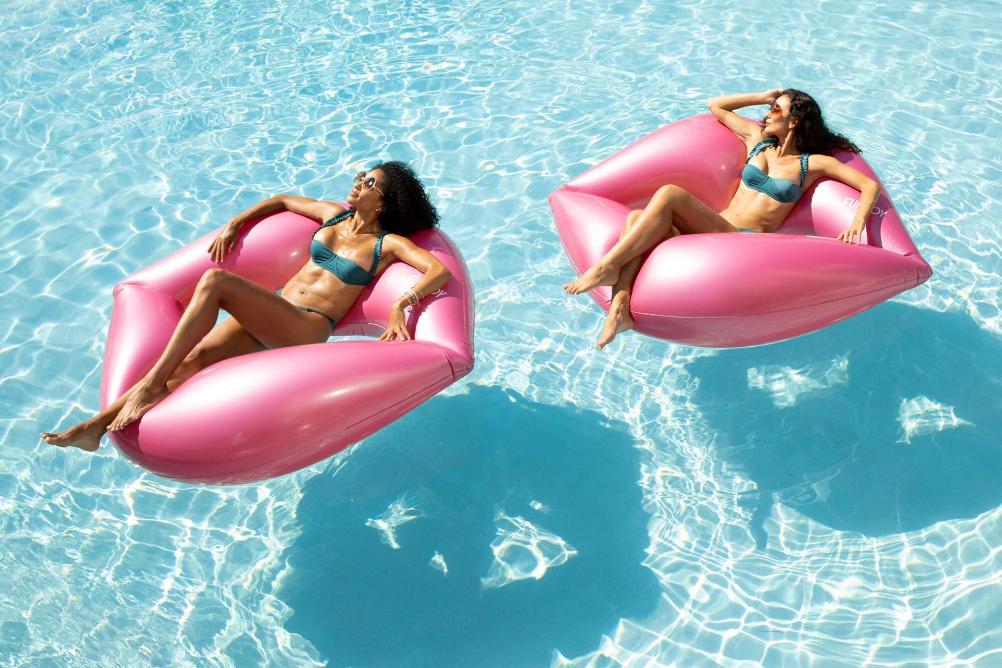 Two individuals floating on pink inflatable tubes in a clear blue pool.