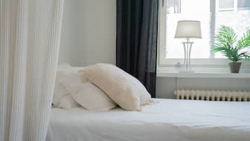 A neatly made bed with white bedding and pillows, a bedside lamp, a potted plant, and a window with curtains.