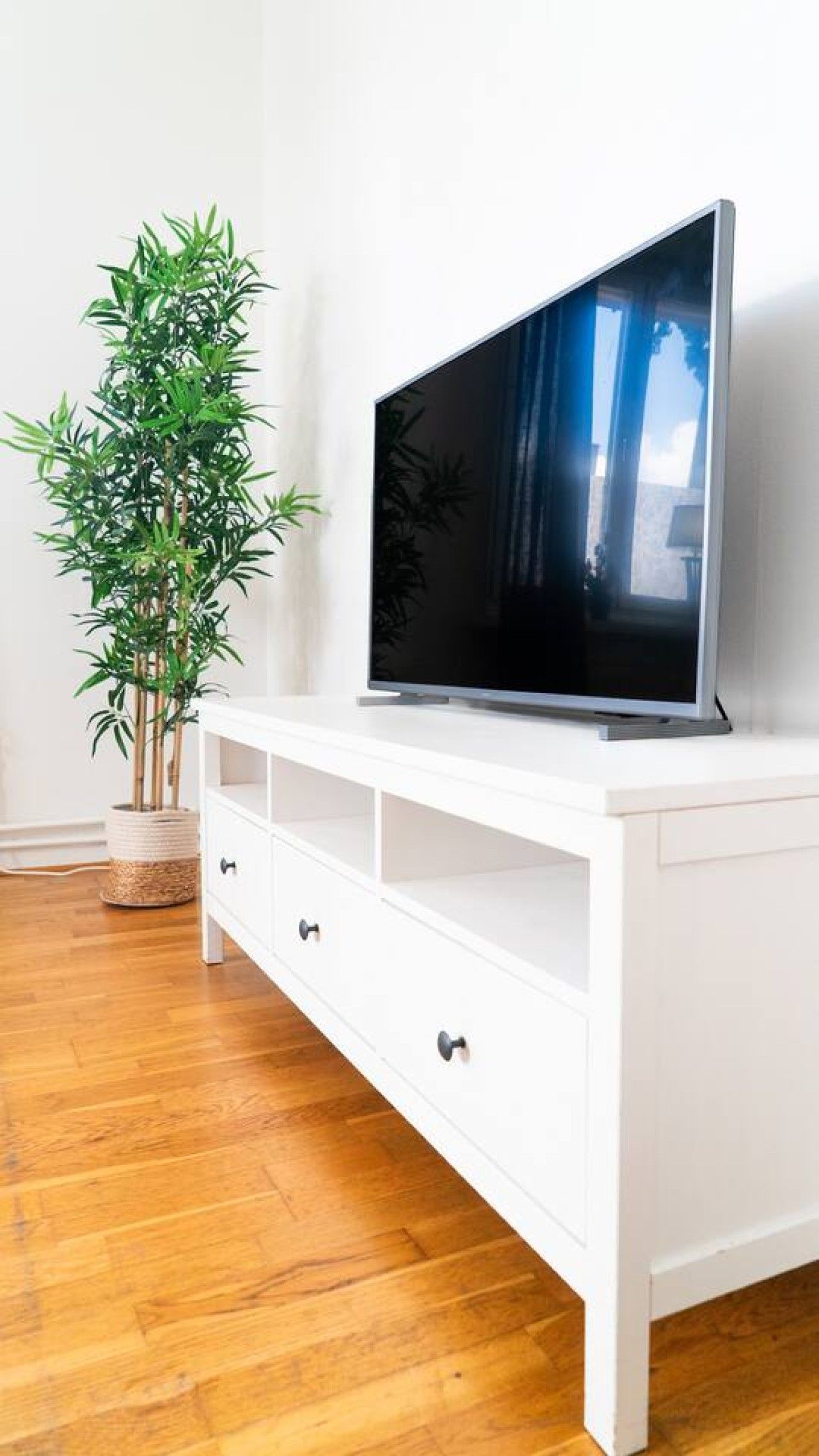 Ett vitt TV-möblemang med en platt-TV, en hög, grön växt och en flätad korg på golvet.