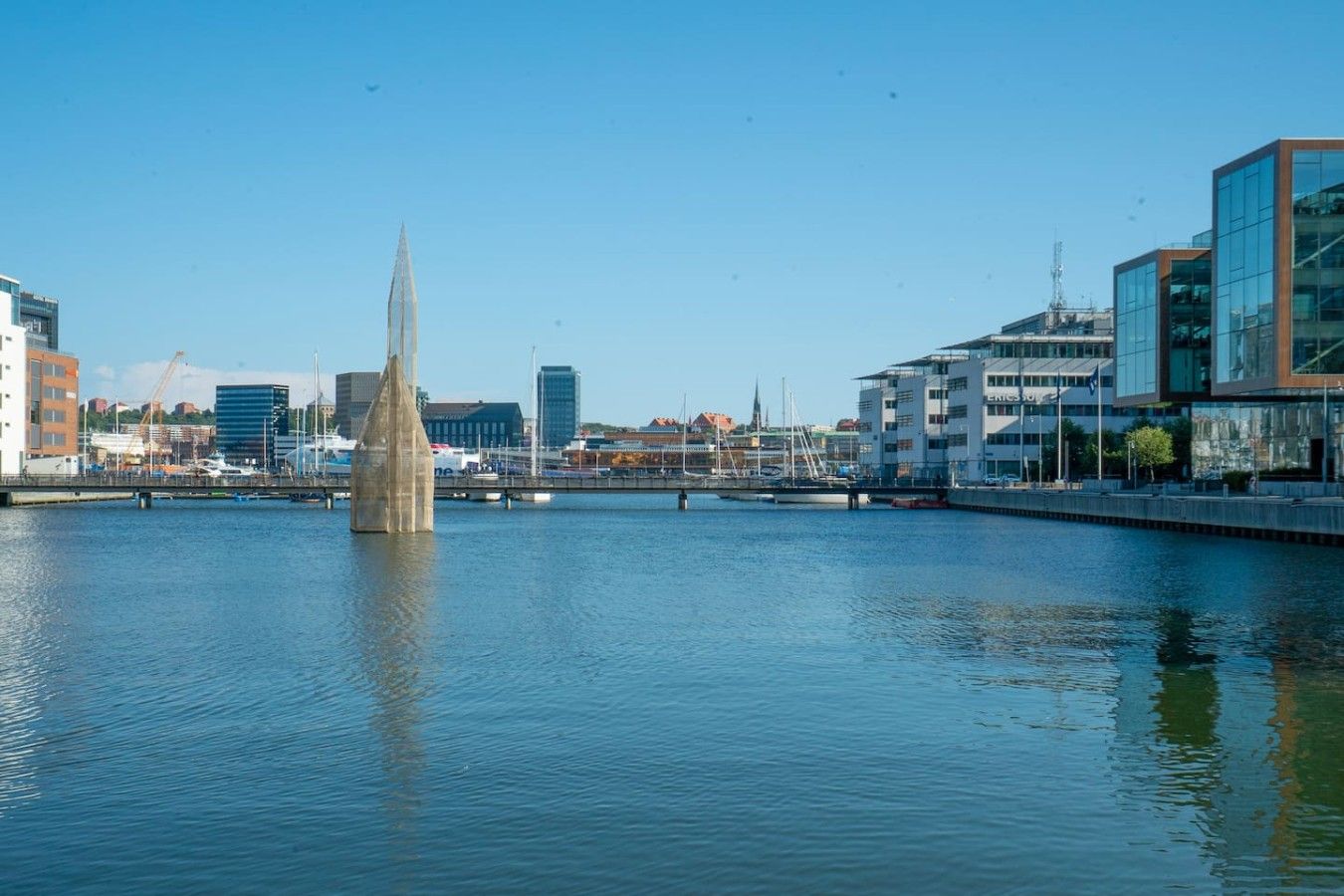 A waterfront view with a tall, slender monument in the center, modern buildings on the right, and a calm body of water reflecting the structures.
