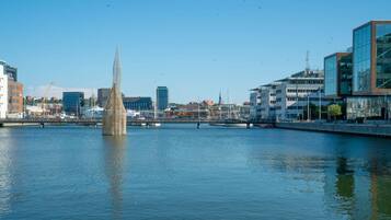 A waterfront view with a tall, slender monument in the center, modern buildings on the right, and a calm body of water reflecting the structures.