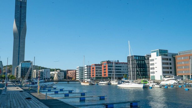 En småbåtshamn med båtar, en hög, modern byggnad och en mängd andra byggnader i bakgrunden.