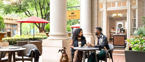 A couple dining outdoors at a cafe with a dog, surrounded by wicker furniture and a large column.
