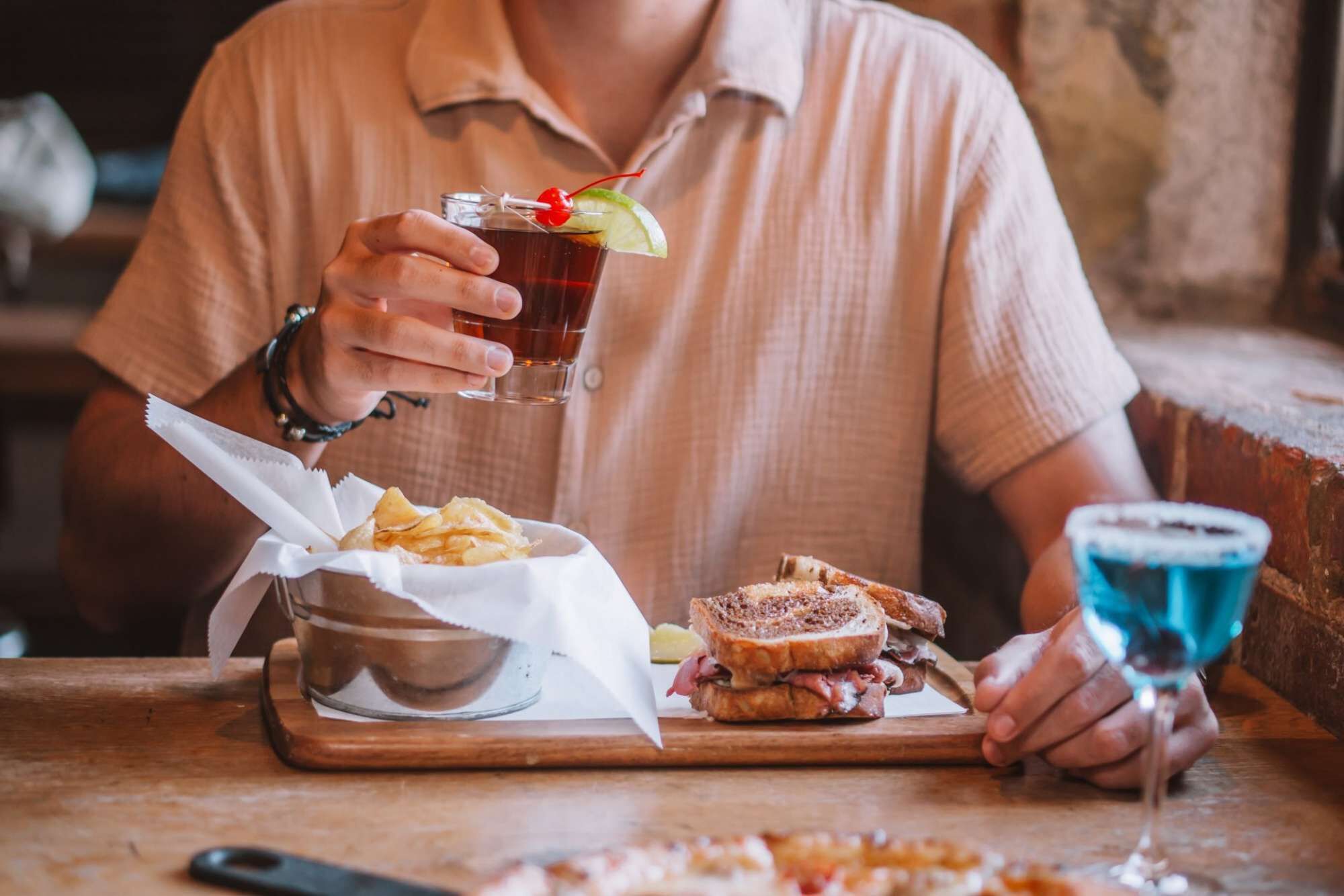 Une personne tenant un verre décoré d’une cerise, assise à une table en bois devant un sandwich et des chips.