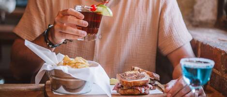 Une personne tenant un verre décoré d’une cerise, assise à une table en bois devant un sandwich et des chips.